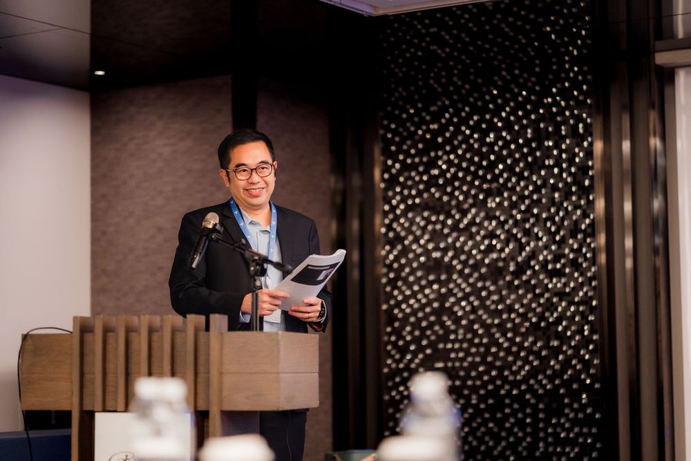 Professional speaking at a corporate event in a modern conference room with decorative wall patterns.