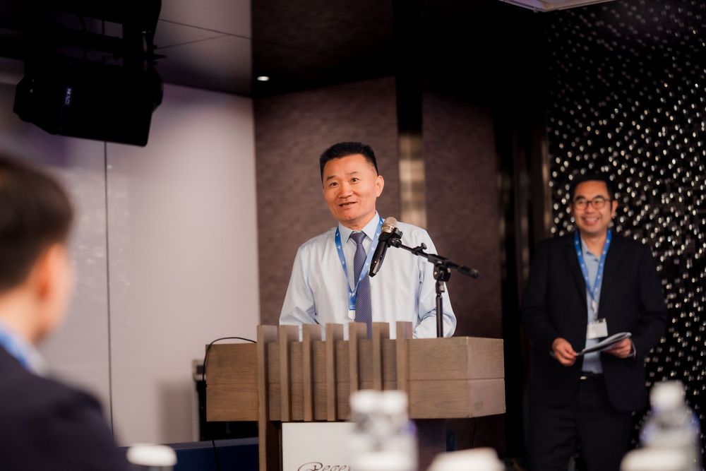 A speaker presents at a business conference podium with attendees in a modern meeting room setting.