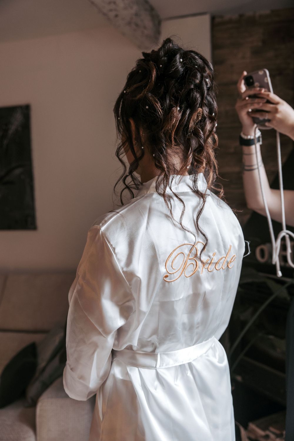 Person in white bridal robe with curled hair styled up takes selfie in mirror during wedding preparations.
