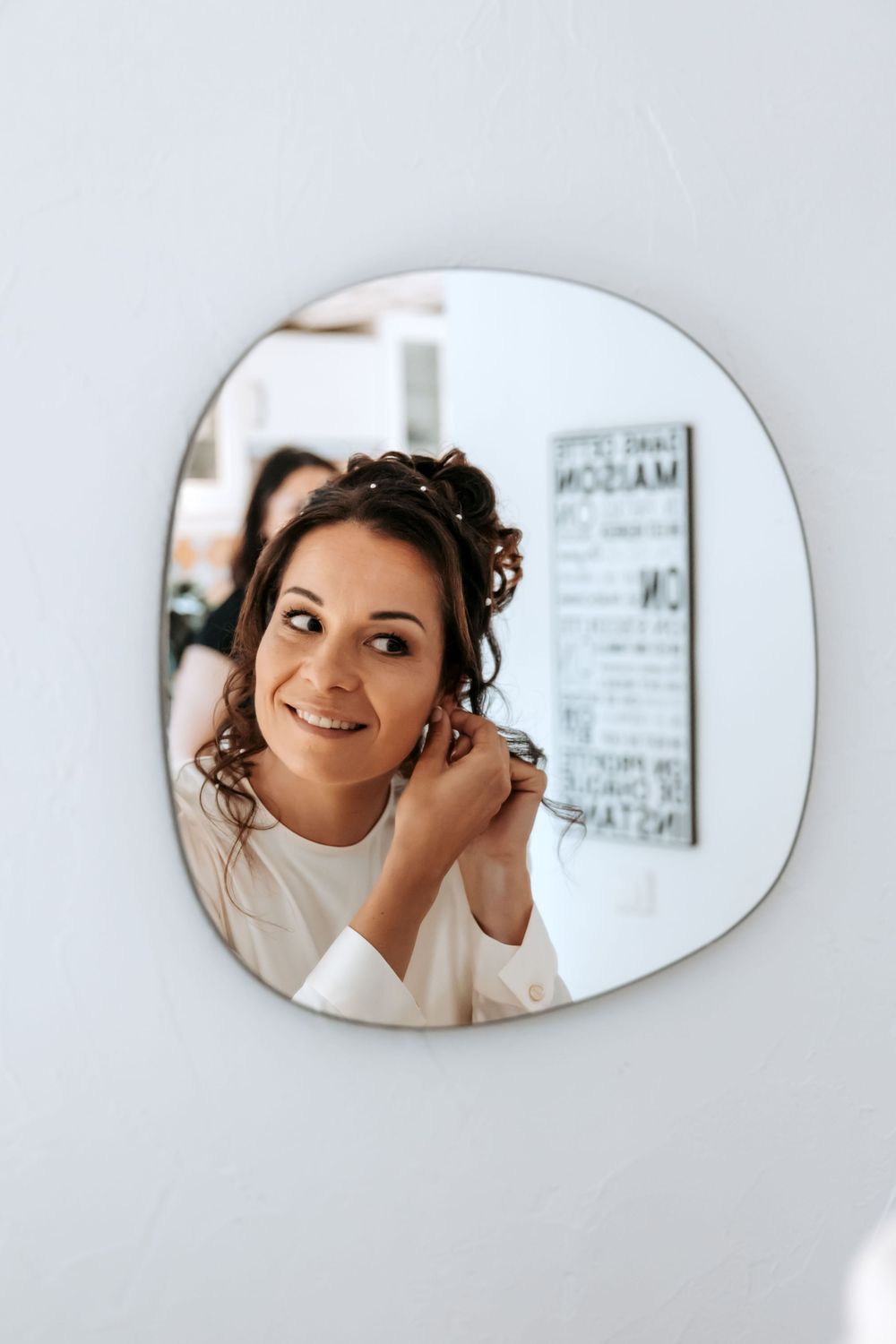 Mirror reflection showing someone putting on earrings while getting ready with a white wall and text sign in background.