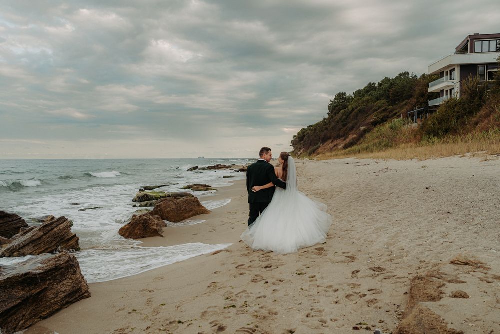 A couple in wedding attire stands together on a rocky beach with cliffs and houses overlooking the ocean on a cloudy day.