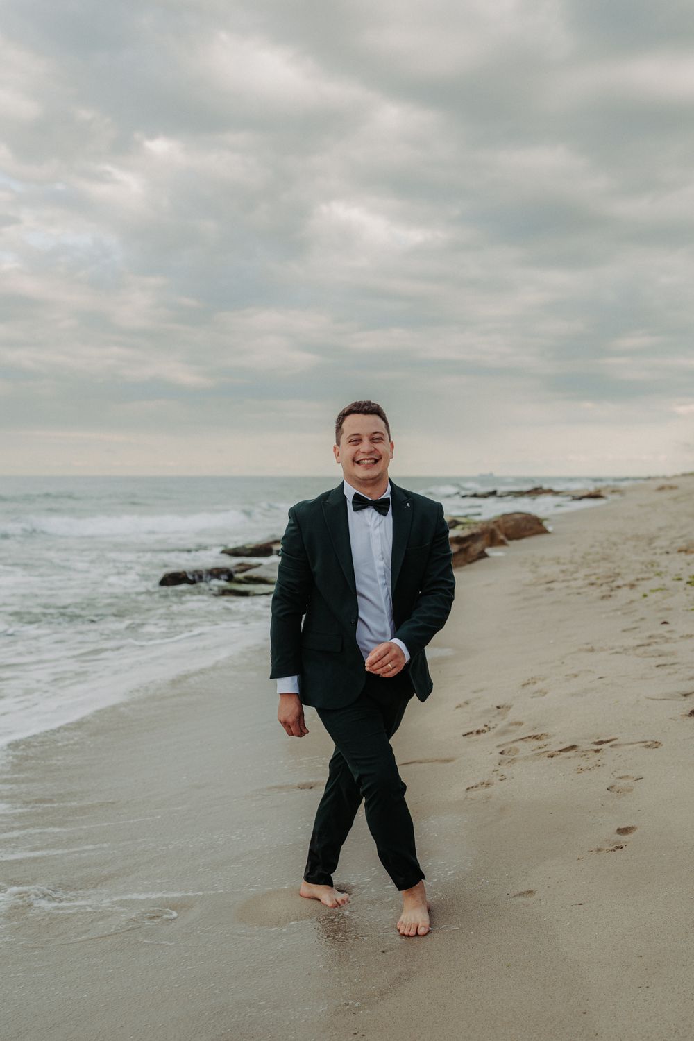 Person in a black tuxedo walking barefoot on a beach with cloudy skies and ocean waves in the background.