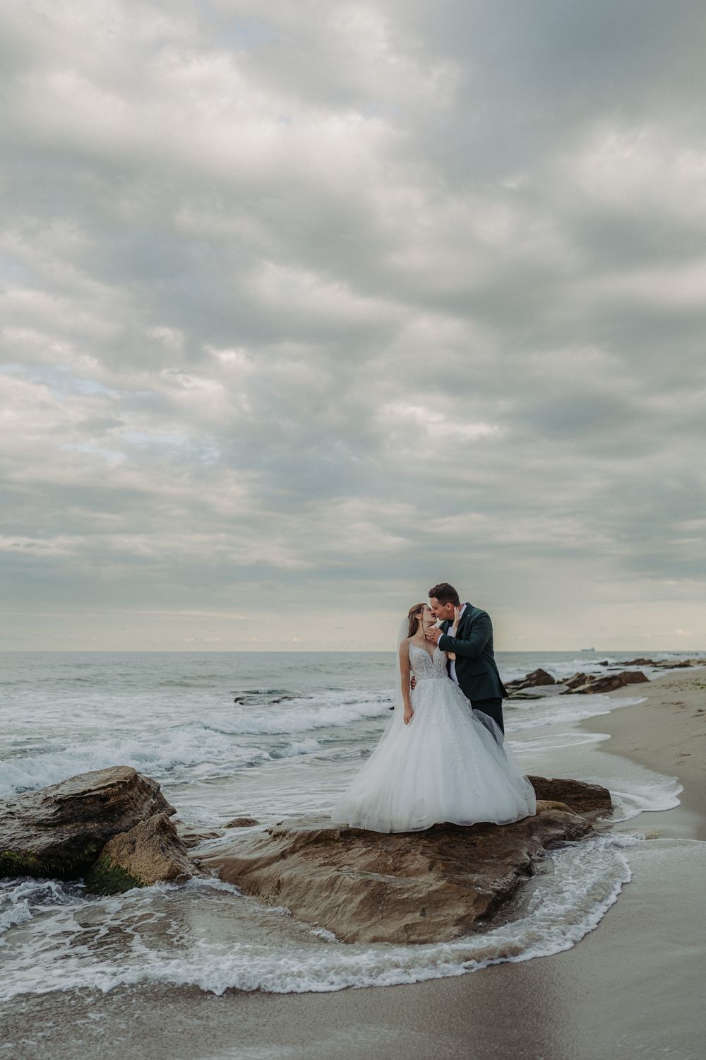A couple in wedding attire sits romantically on coastal rocks as waves crash around them at the beach.