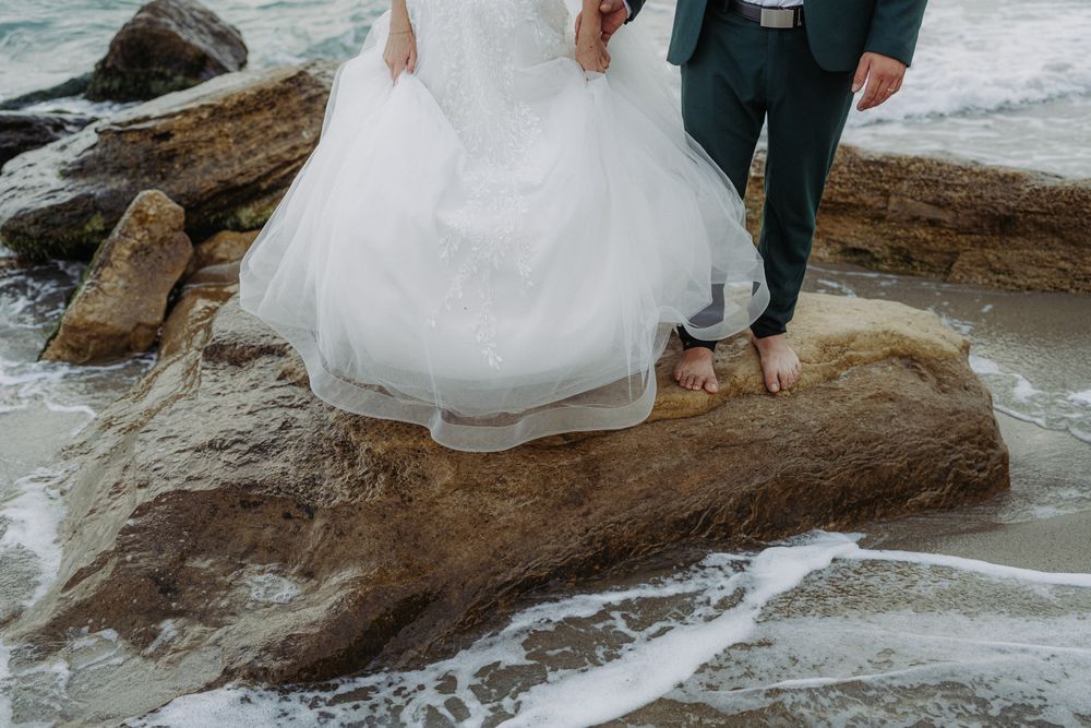 A couple in wedding attire poses on rocky coastal cliffs with ocean waves crashing in the background.
