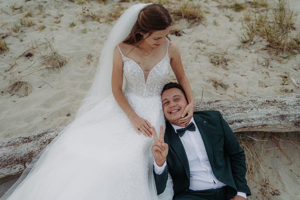 A couple in wedding attire poses together on a sandy beach slope.