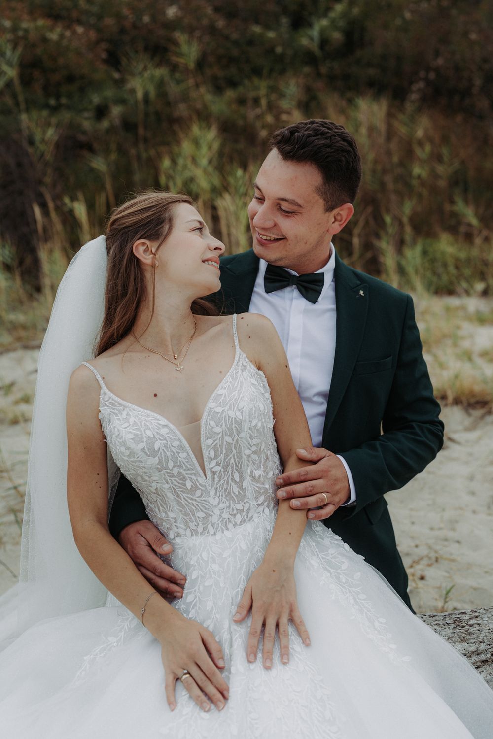 Newlyweds in formal attire share loving moments on a beach with the bride in a white lace gown.