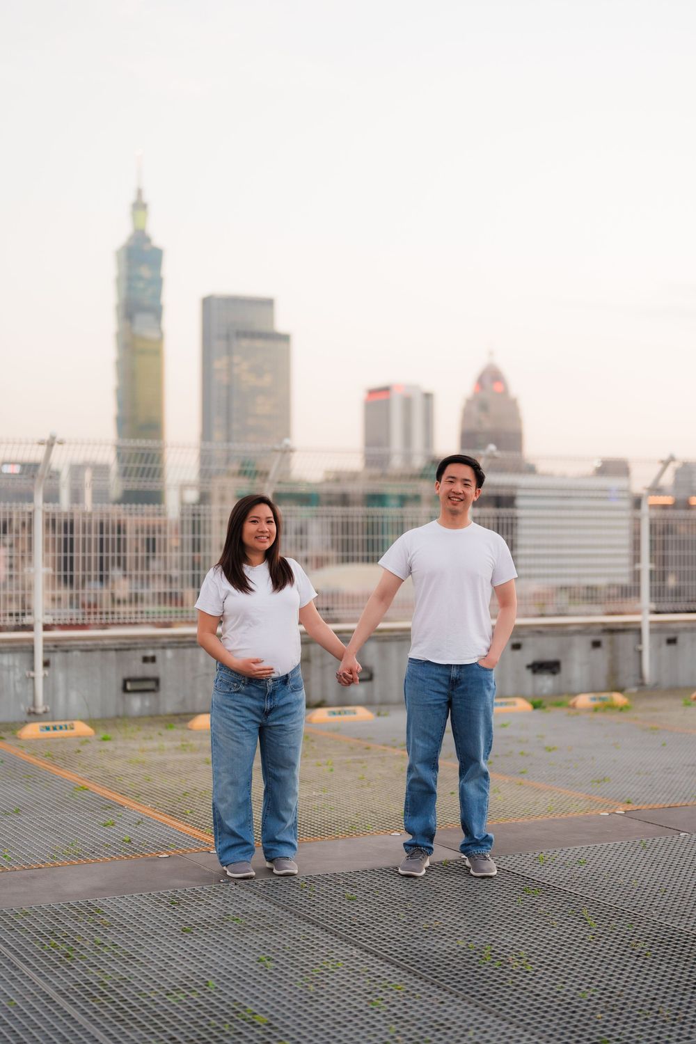 Couple in matching white shirts and jeans holds hands on urban rooftop with city skyline backdrop.