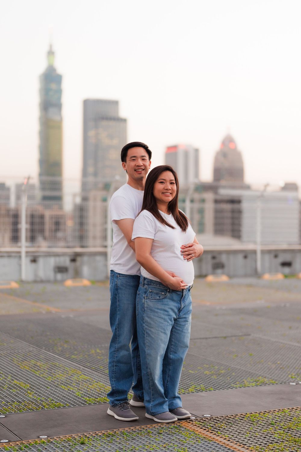 Expectant couple poses on rooftop with Taipei 101 and city skyline in background during sunset.