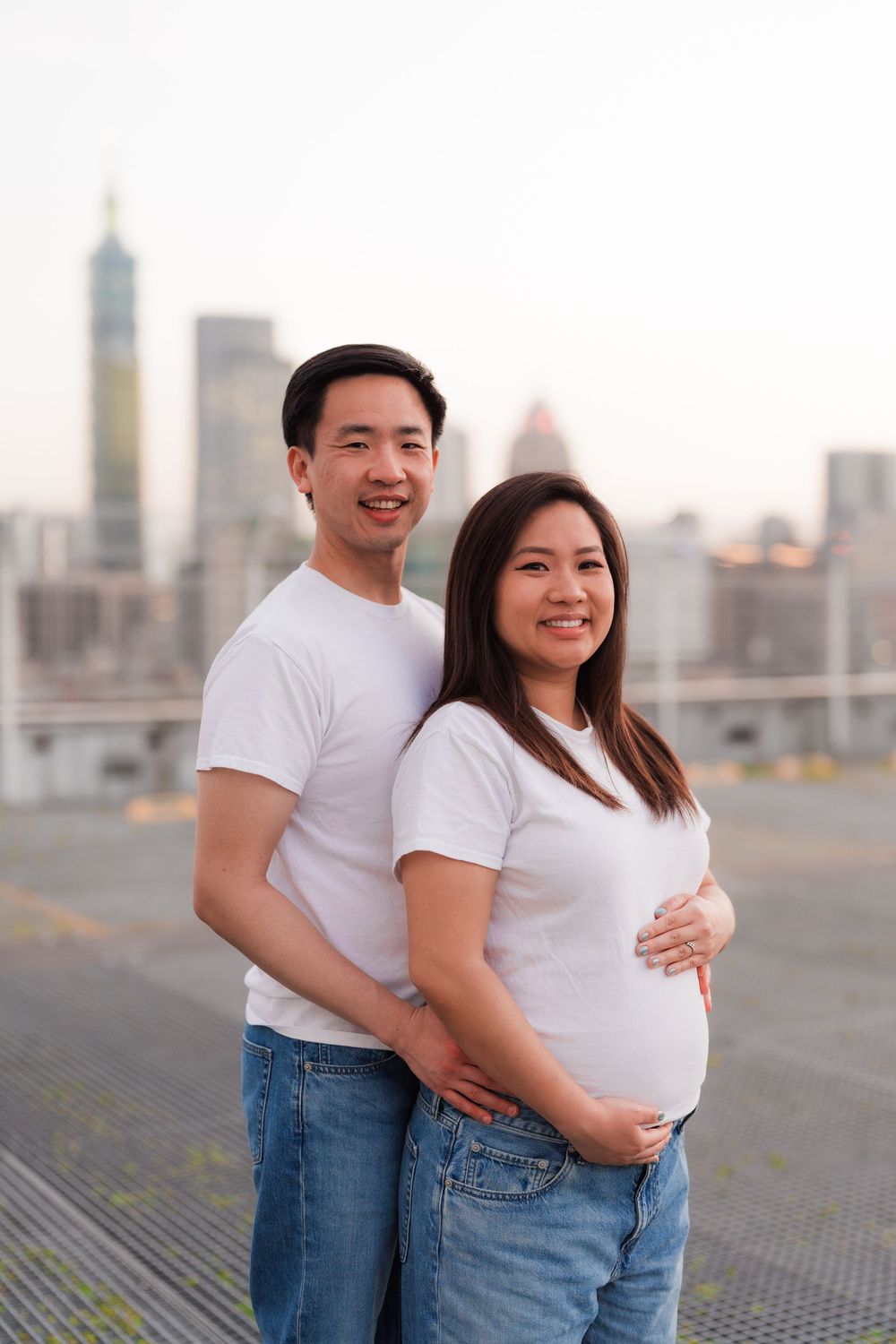 Maternity photoshoot on rooftop features couple in casual white shirts with cityscape view.