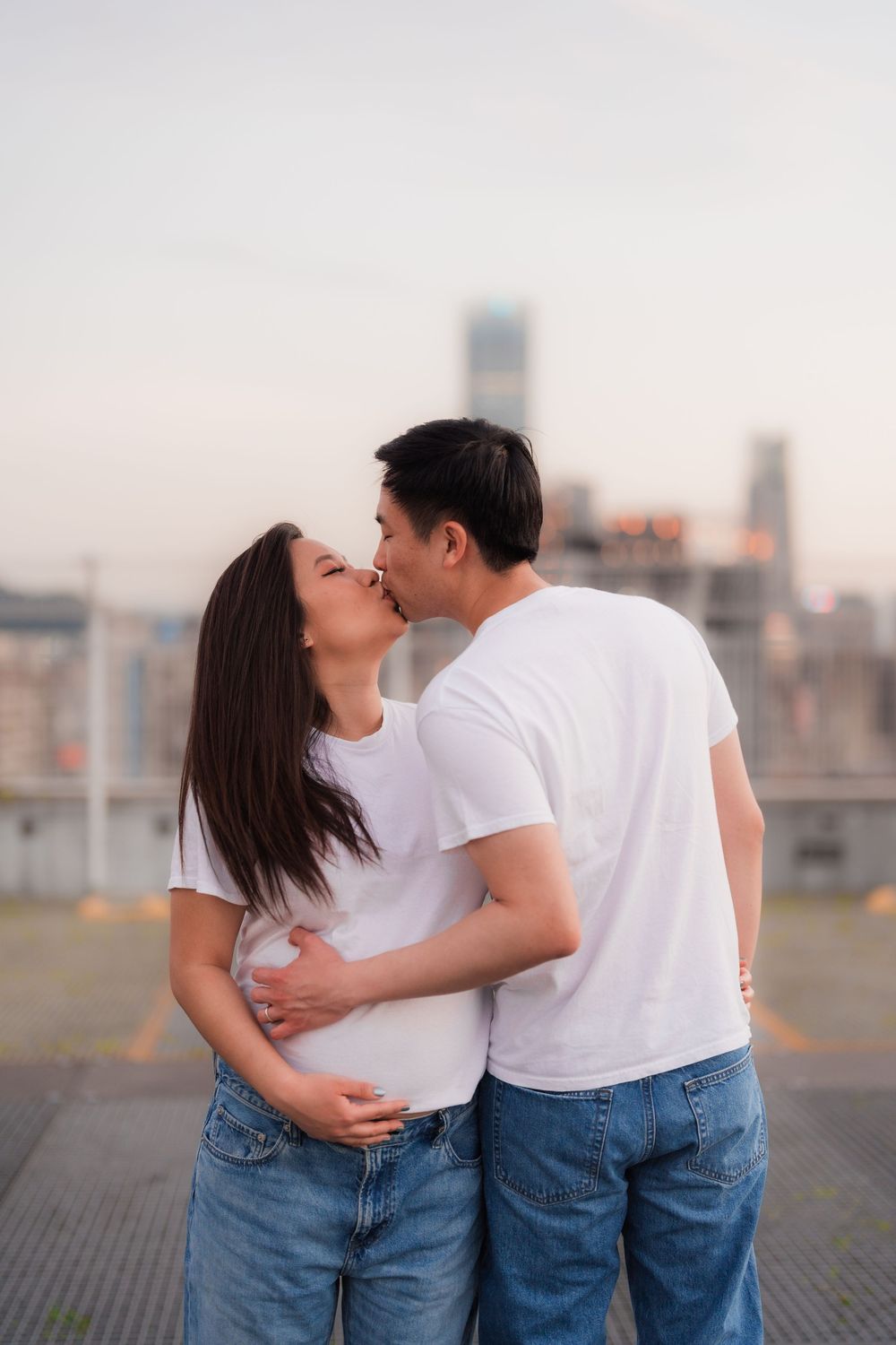 A couple in matching white t-shirts and jeans share a romantic moment on a rooftop with a city skyline backdrop.