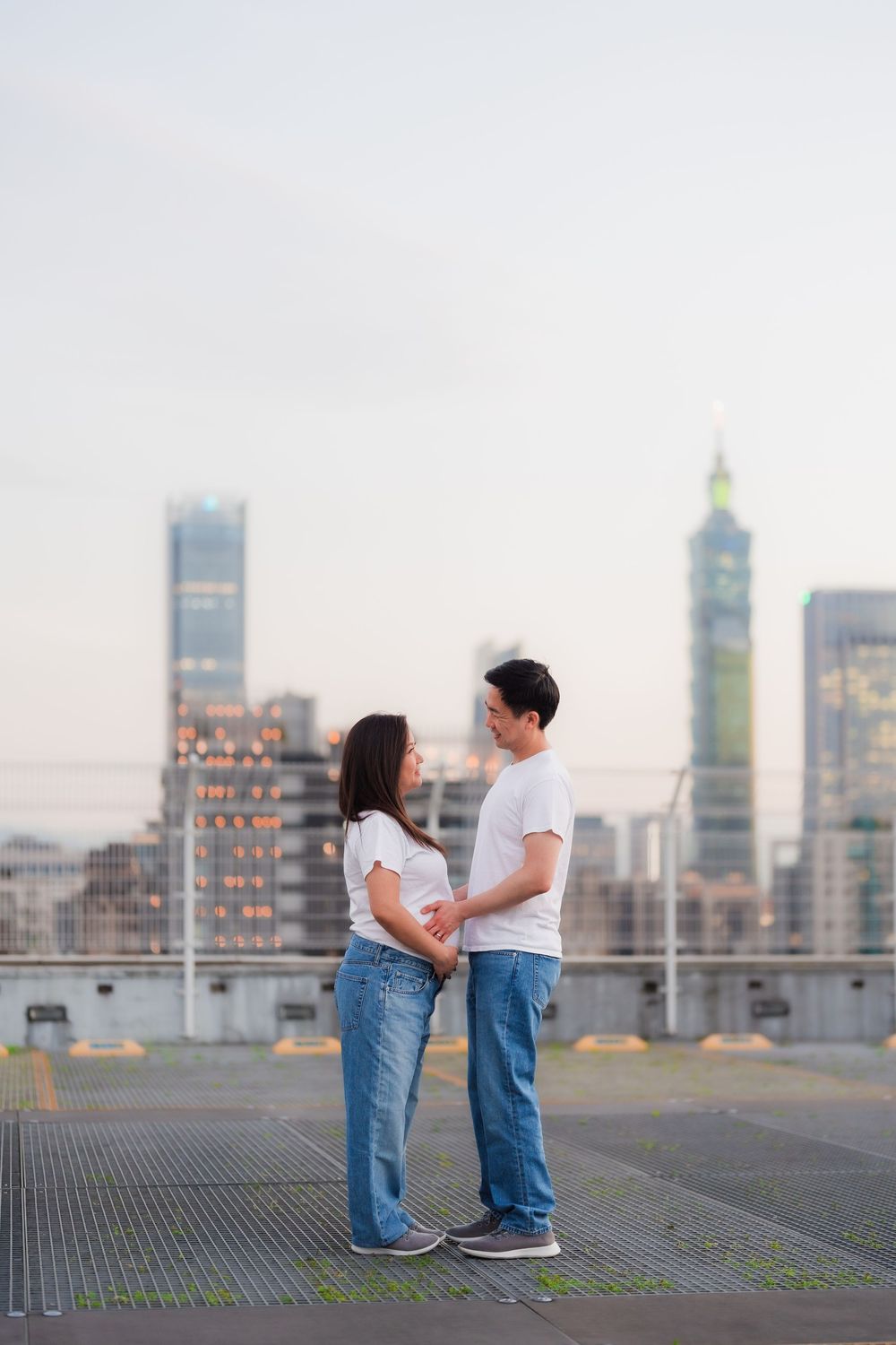 A couple in matching white shirts and jeans pose against a cityscape backdrop at sunset.