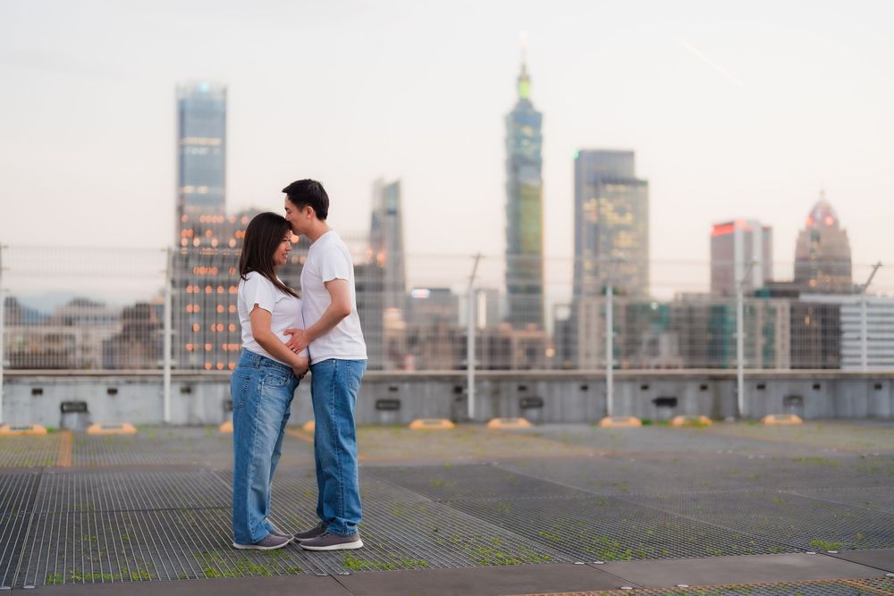 Two figures embrace on a rooftop terrace with a cityscape of tall buildings and skyscrapers in the background at dusk.