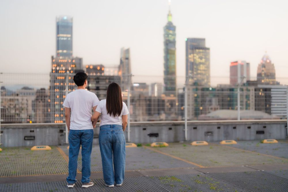 A couple in casual outfits stands together overlooking an urban skyline at dusk with illuminated buildings.