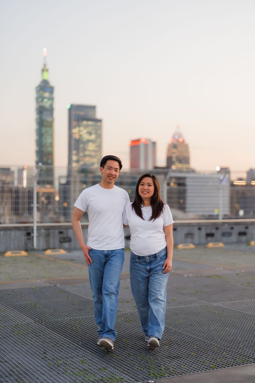 A couple in matching white t-shirts and jeans pose on a rooftop with a city skyline at sunset.