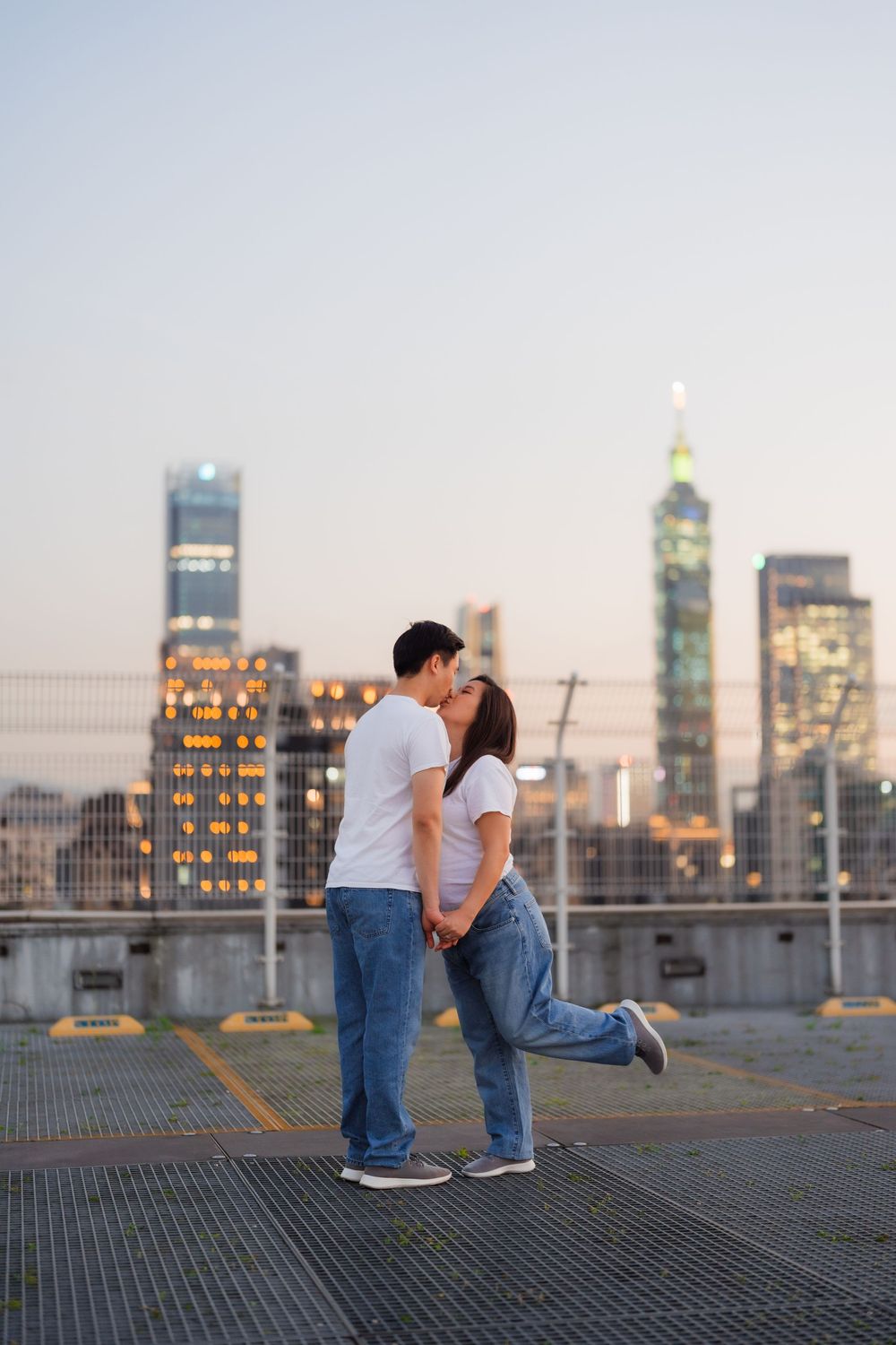 A romantic couple shares a kiss on an urban rooftop with Taipei City skyline skyscrapers in the background at dusk.