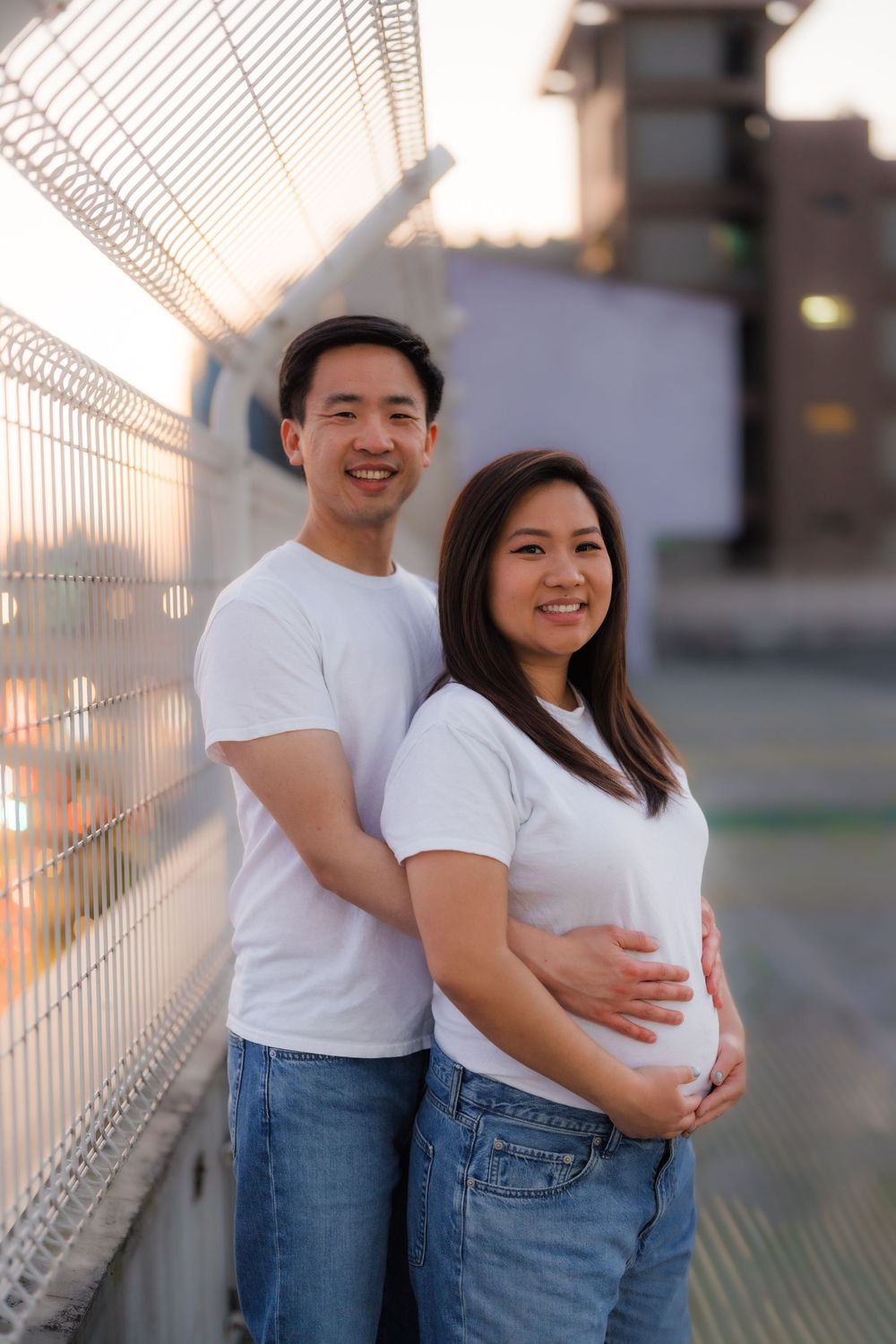 A man embraces his pregnant wife at sunset standing near the edge of a tall skyscraper in matching white shirts.
