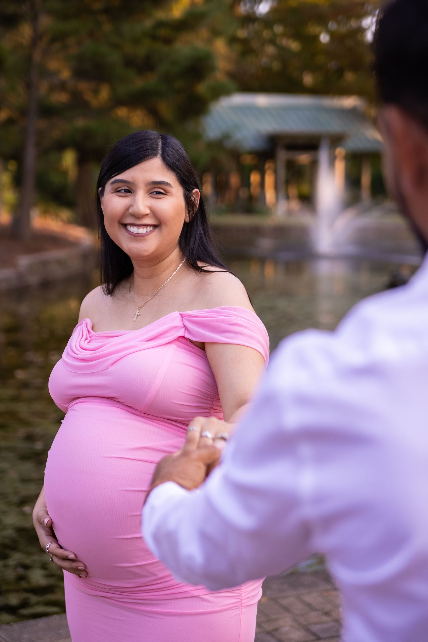Maternity photo session by fountain with woman in pink off-shoulder dress during golden hour at Mercer Botanic Garden in Houston Tx.
