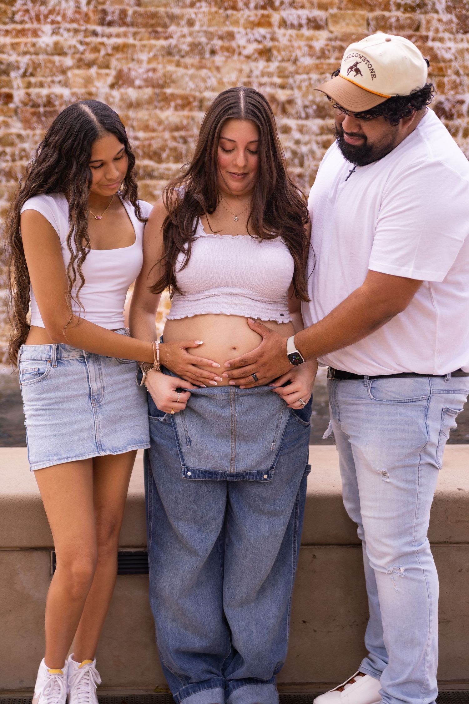 Casual pregnancy announcement photos against brick wall wearing white tops and denim by the water way in the Woodlands TX.