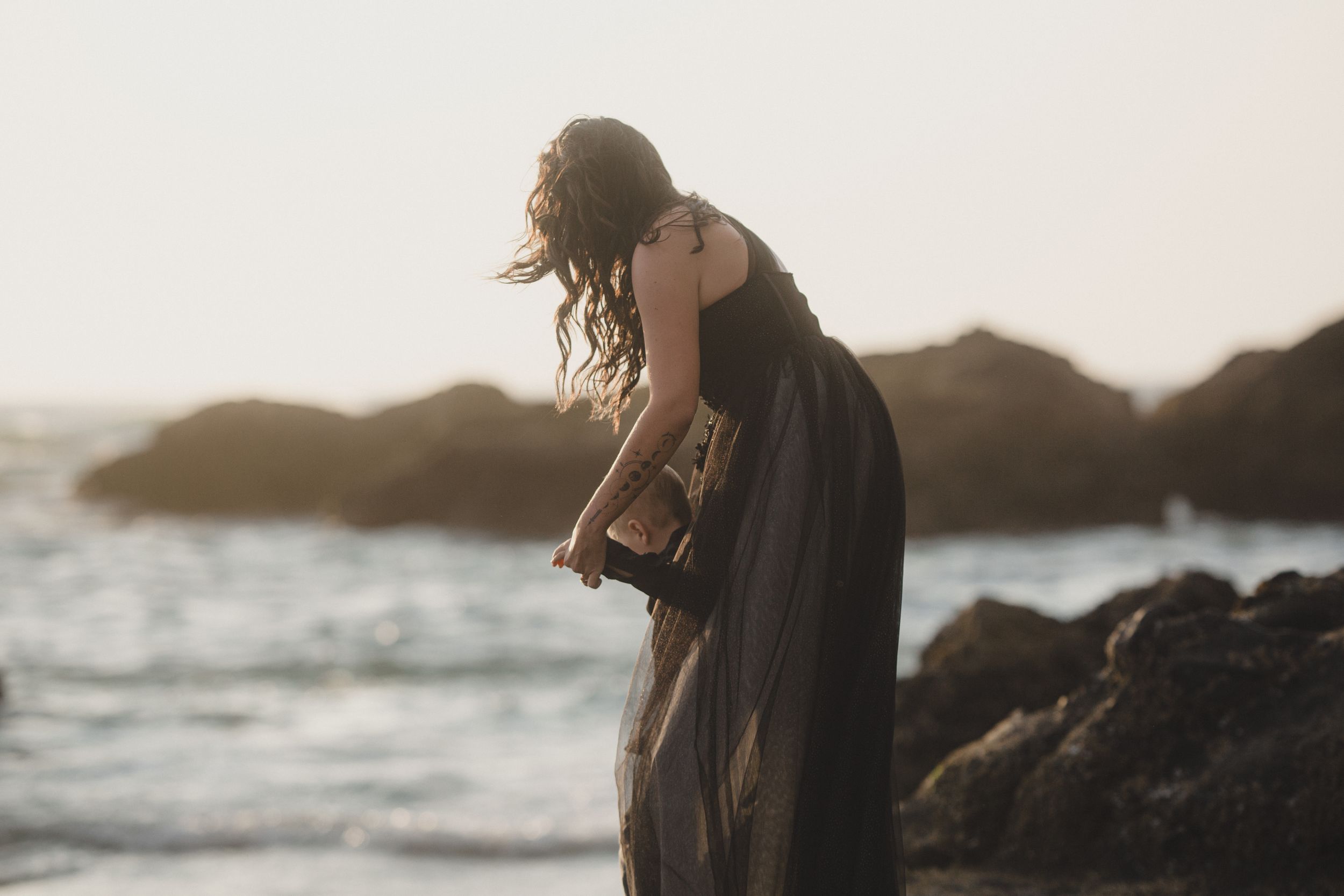 A couple embraces on a rocky beach at sunset with waves crashing in the background.