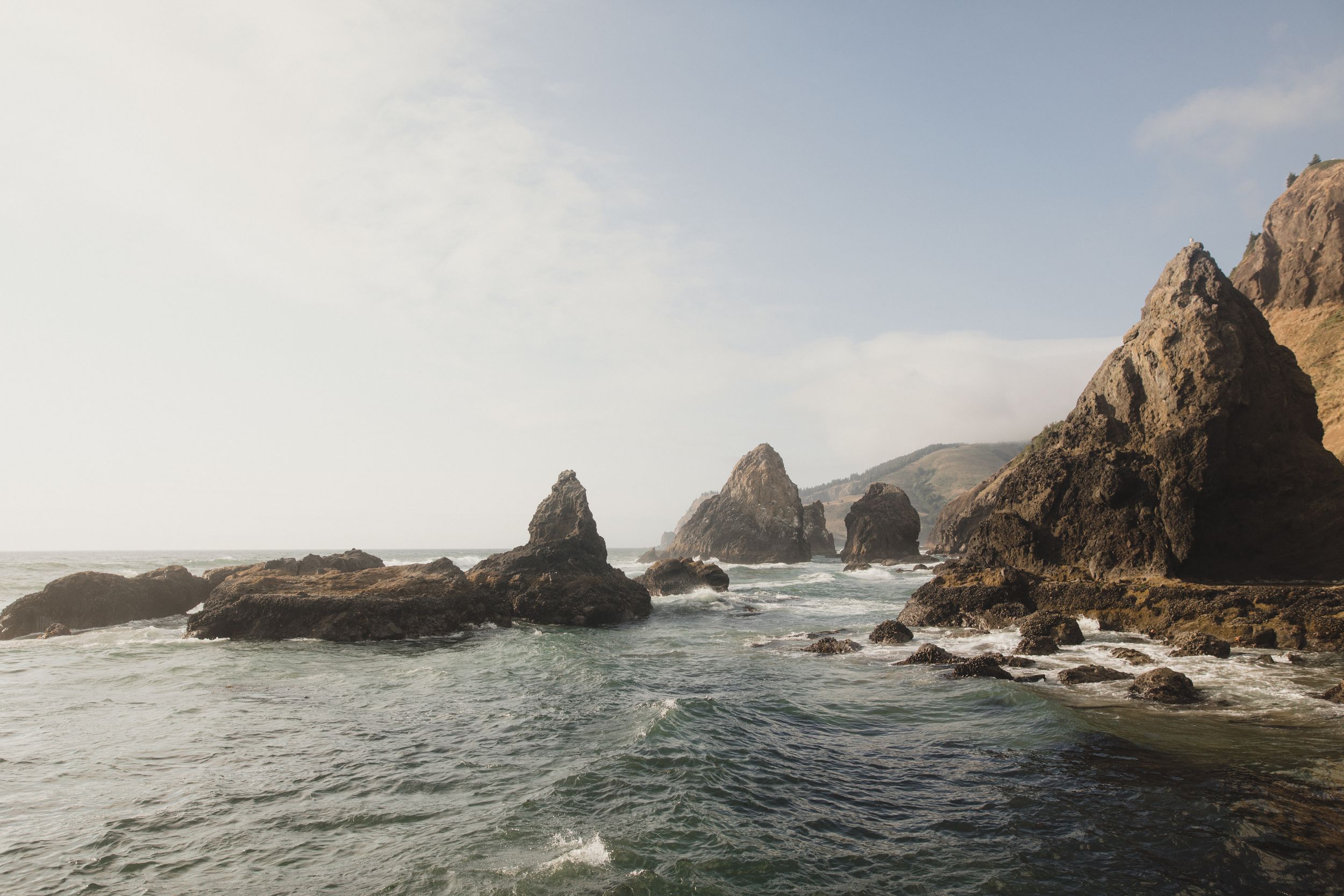 Dramatic rocky coastline with waves crashing against rugged cliffs and sea stacks along the Pacific Ocean shore.