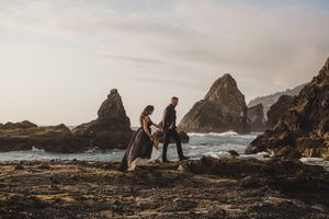 Two people walking on rocky coastal shore with dramatic sea stacks and crashing waves at sunset.
