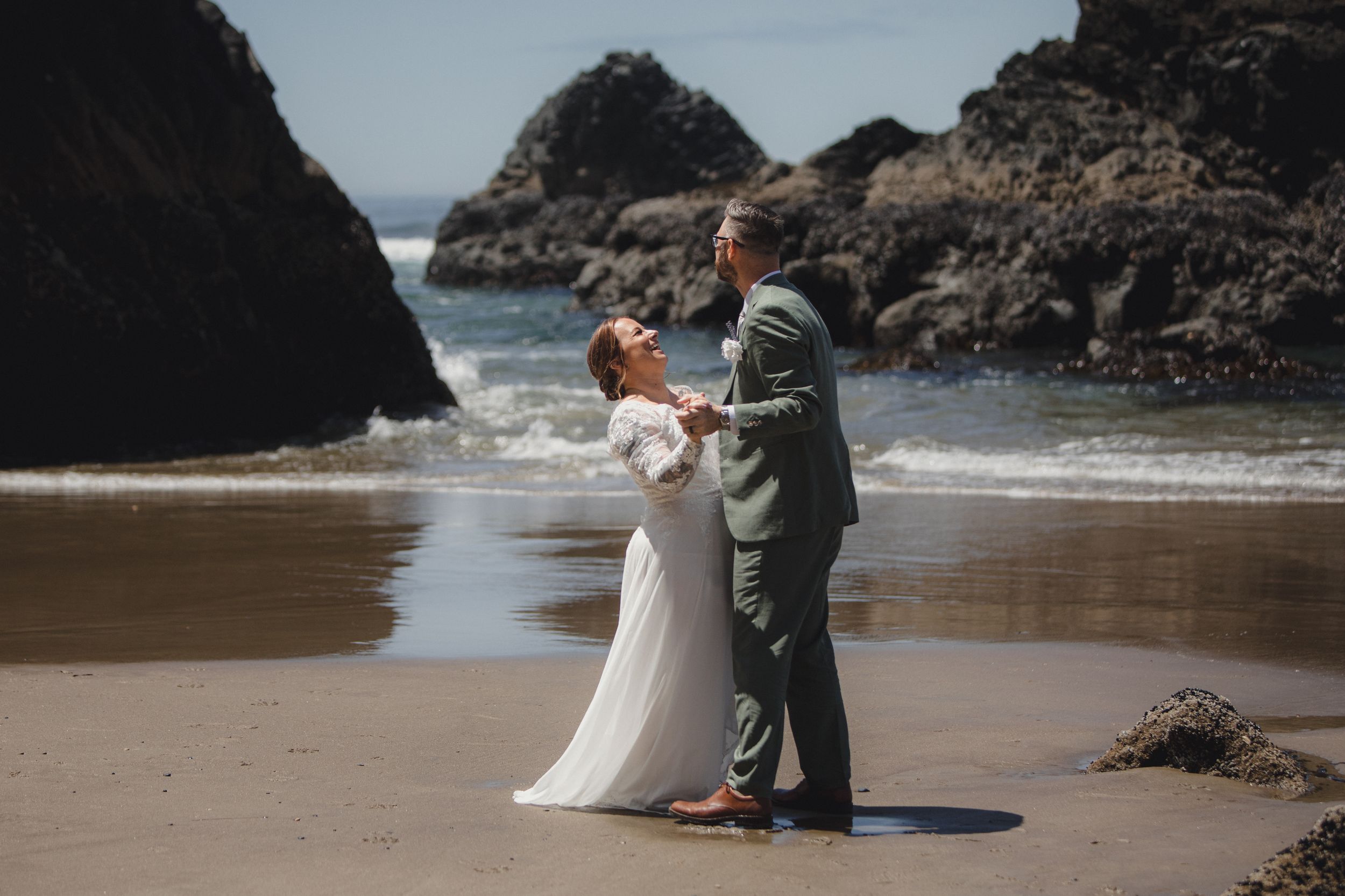 A couple in formal wear embraces on a scenic beach surrounded by dramatic rock formations and ocean waves.
