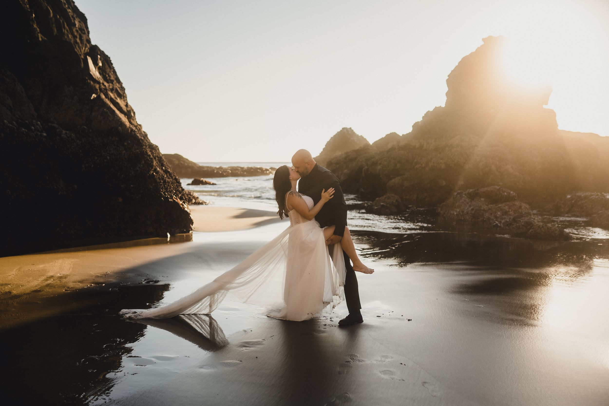 Two silhouettes embrace on a rocky beach at sunset as golden light streams through coastal cliffs.