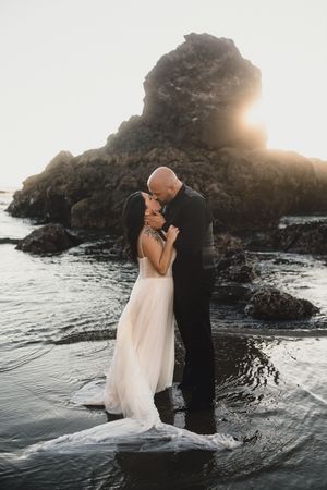 Couple shares romantic sunset moment in wedding attire along rocky coastal shoreline with crashing waves and dramatic lighting.