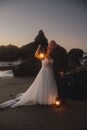 A romantic beach wedding photo session at sunset with glowing lanterns and rocky coastal backdrop.