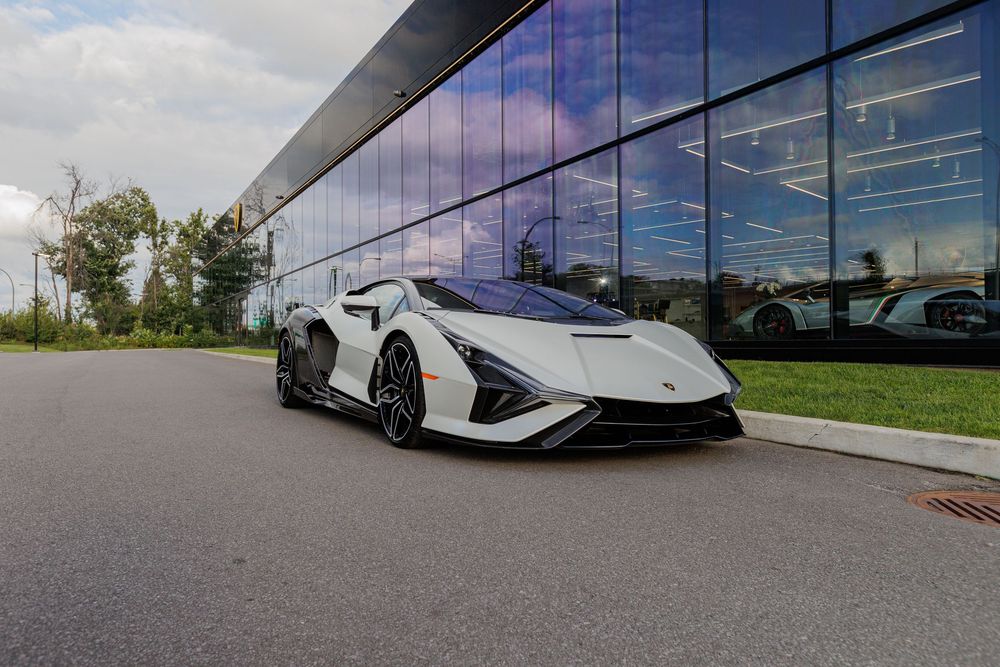A white Lamborghini Sian supercar parked in front of a modern glass building with reflective windows on a cloudy day.