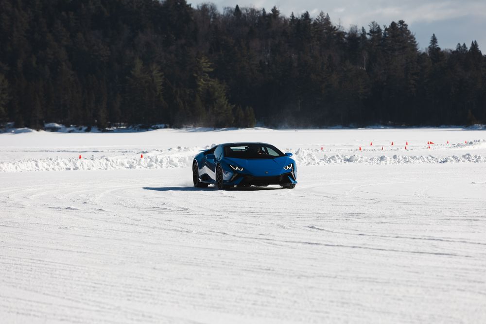 A blue car drives across a vast snowy frozen lake surrounded by evergreen forest on a winter day.