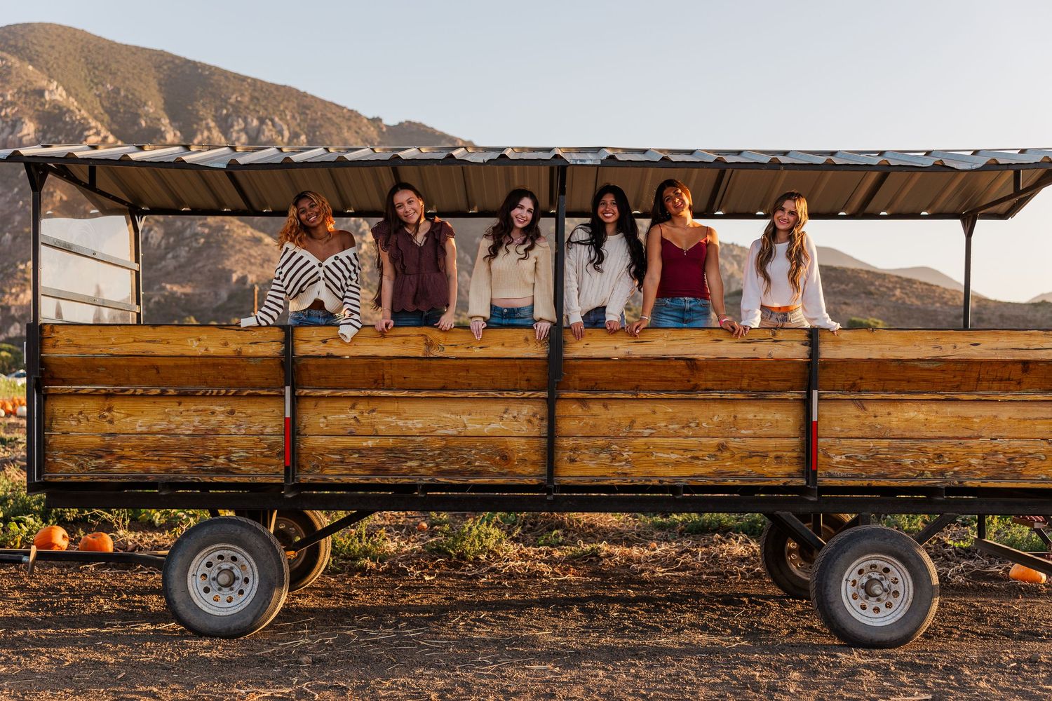 A group of people sit together on a wooden hay wagon in front of desert mountains during sunset.