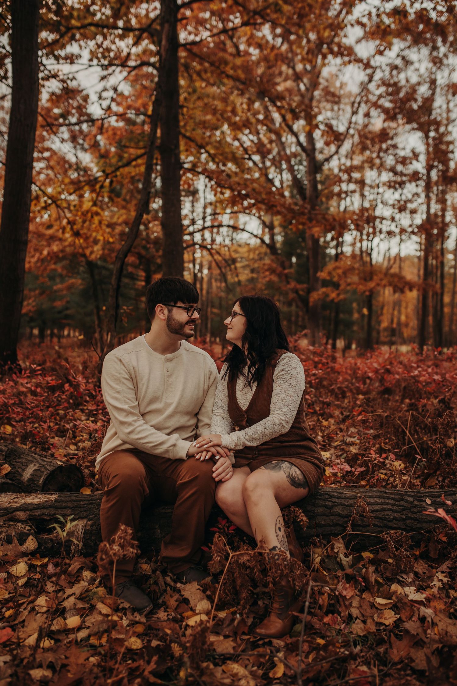 A couple shares intimate moments while sitting among fallen autumn leaves in a warm-toned forest setting.