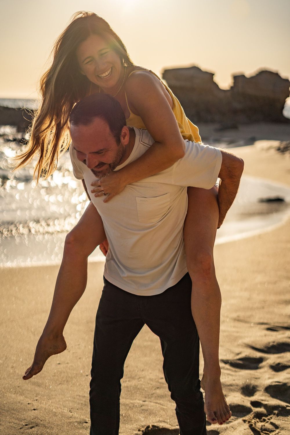 A playful couple shares a piggyback ride on a beach at sunset with ocean waves and rocks in the background.