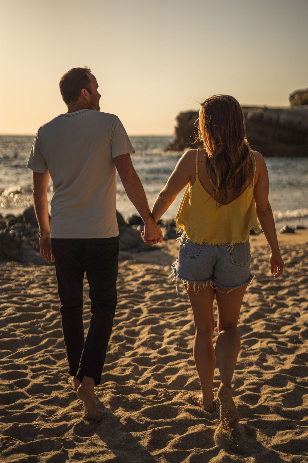 Couple walking hand in hand on a sandy beach at sunset casting long shadows on the sand.