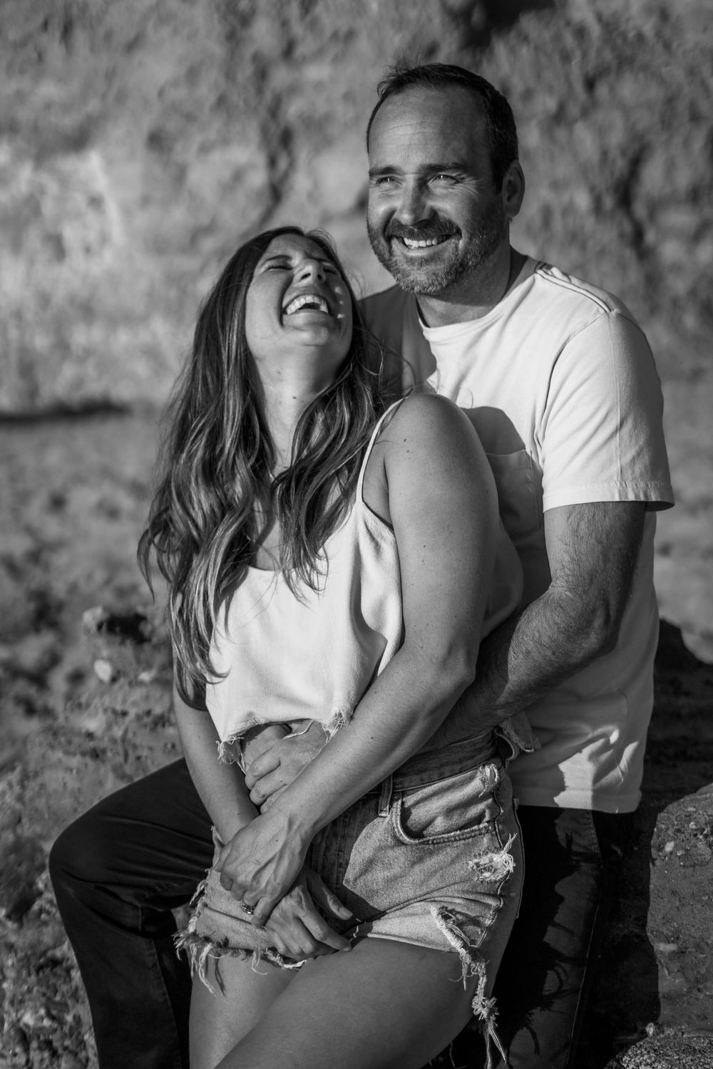 A romantic black and white photo series showing a couple laughing and embracing on a rocky beach.