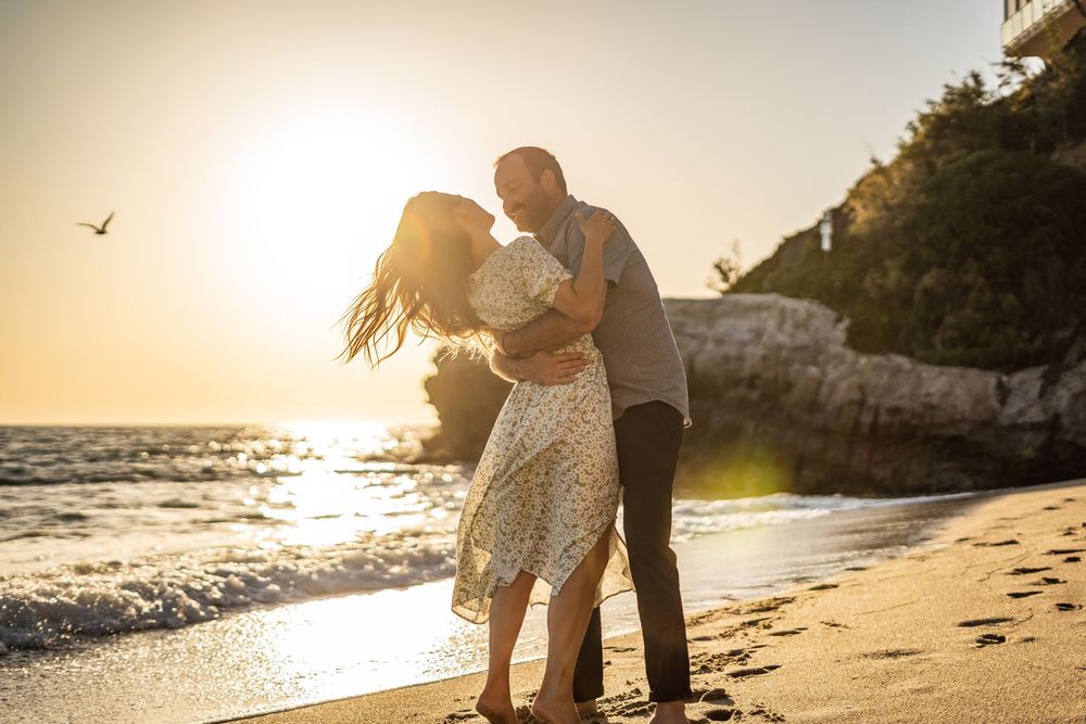 A couple embraces on a golden beach at sunset with waves crashing on the shore and cliffs in the background.