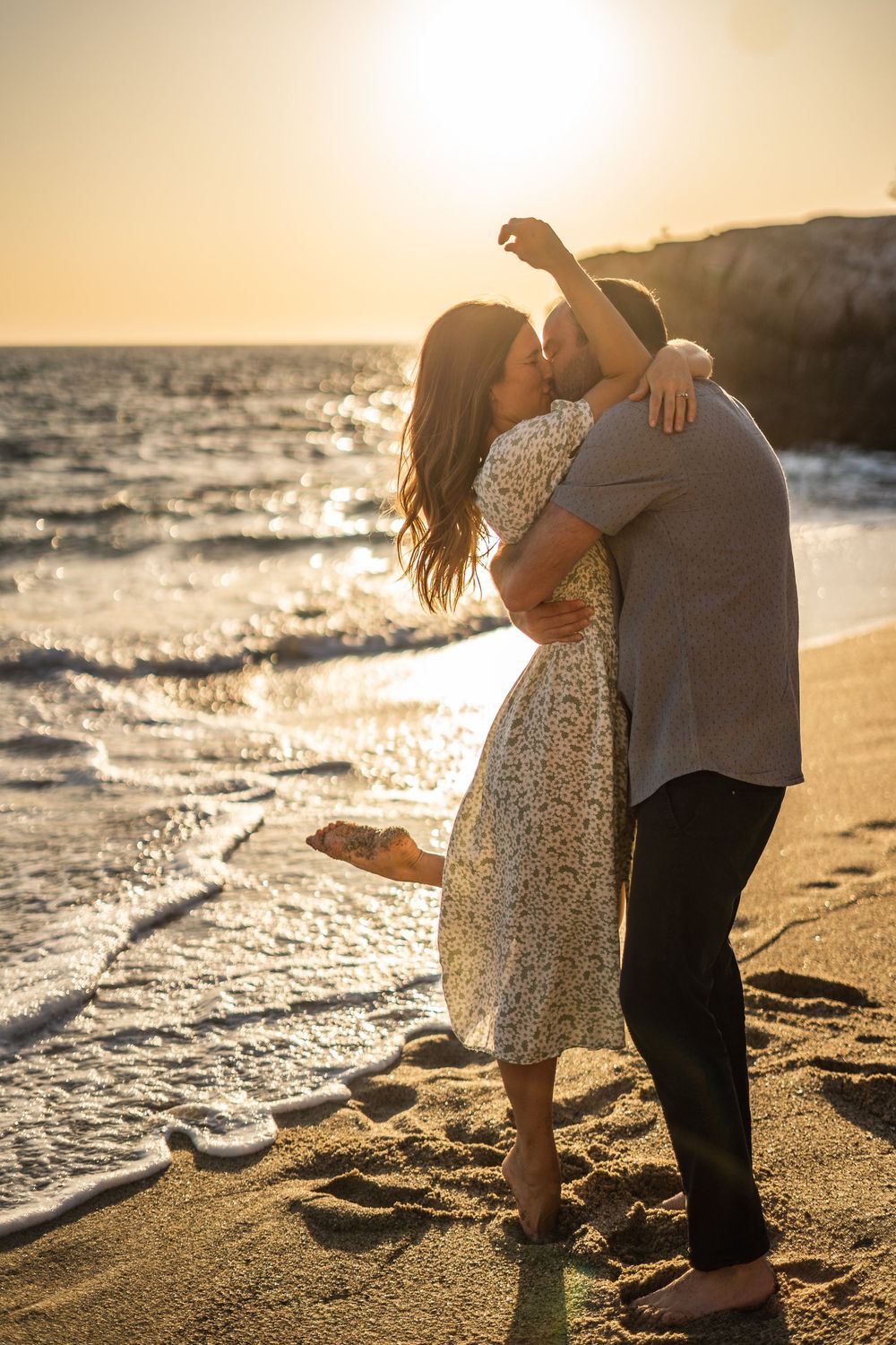 A couple embraces on a golden beach at sunset as waves crash on the shore, creating a romantic silhouette.