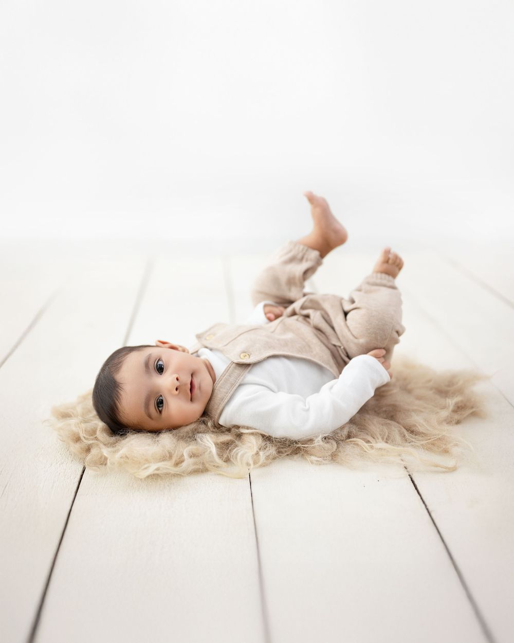 Baby in white outfit lying on light wooden floor during professional photo session.