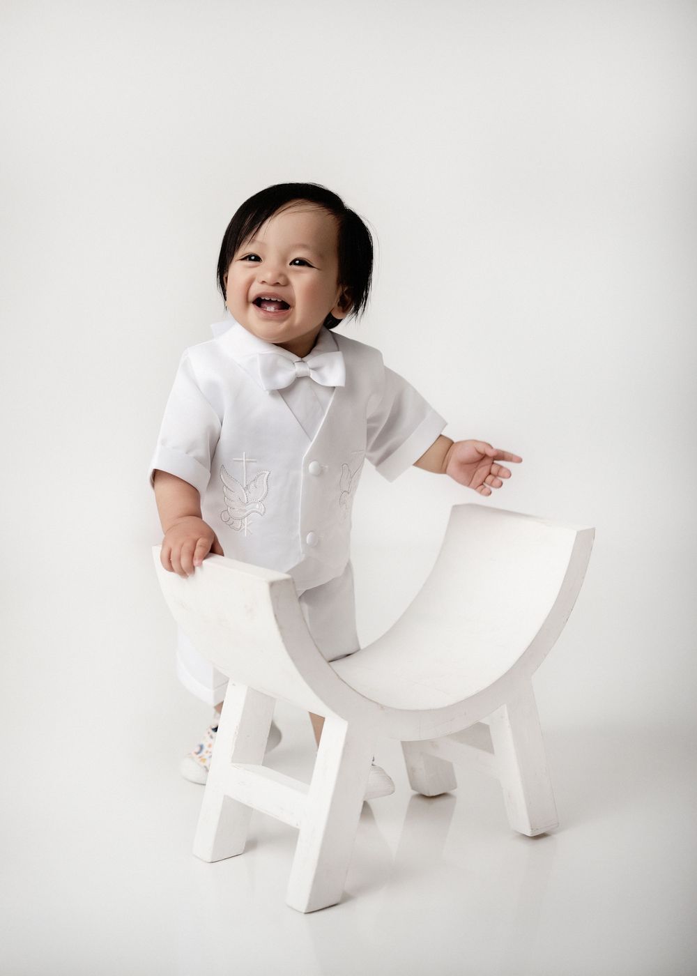 Baby in white outfit sits on decorative white chair during studio photoshoot.