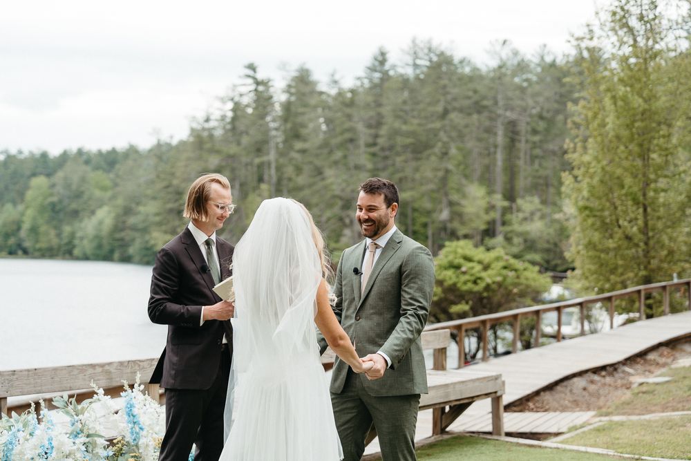 A wedding couple shares a joyful moment together on a rustic wooden dock surrounded by evergreen trees and water. Asheville, NC - Camp