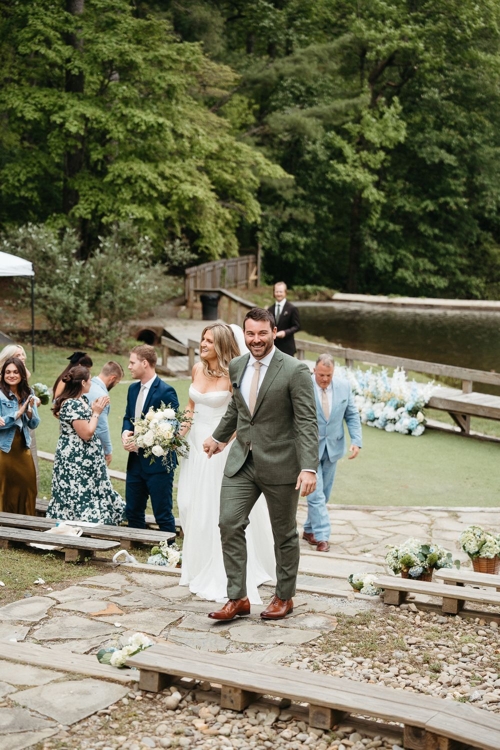 A wedding celebration unfolds at a rustic outdoor venue with guests seated on wooden benches surrounded by lush greenery. Hendersonville, NC