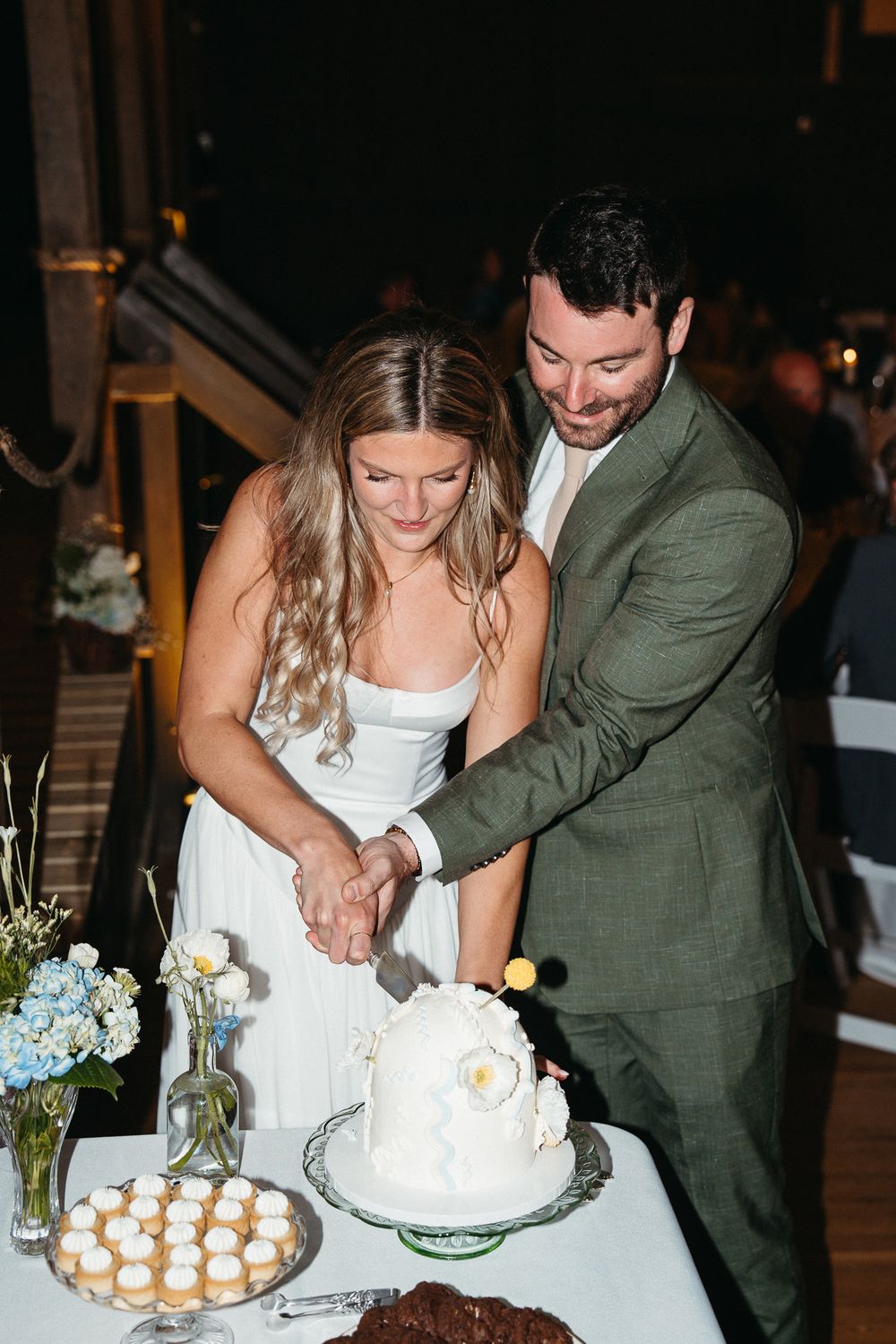 A couple cuts their wedding cake together while guests watch at an evening reception. Asheville, NC