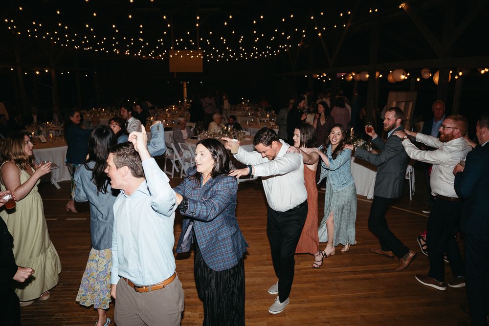 Wedding guests dance in a line formation on a dark wooden dance floor under twinkling lights. Asheville, NC