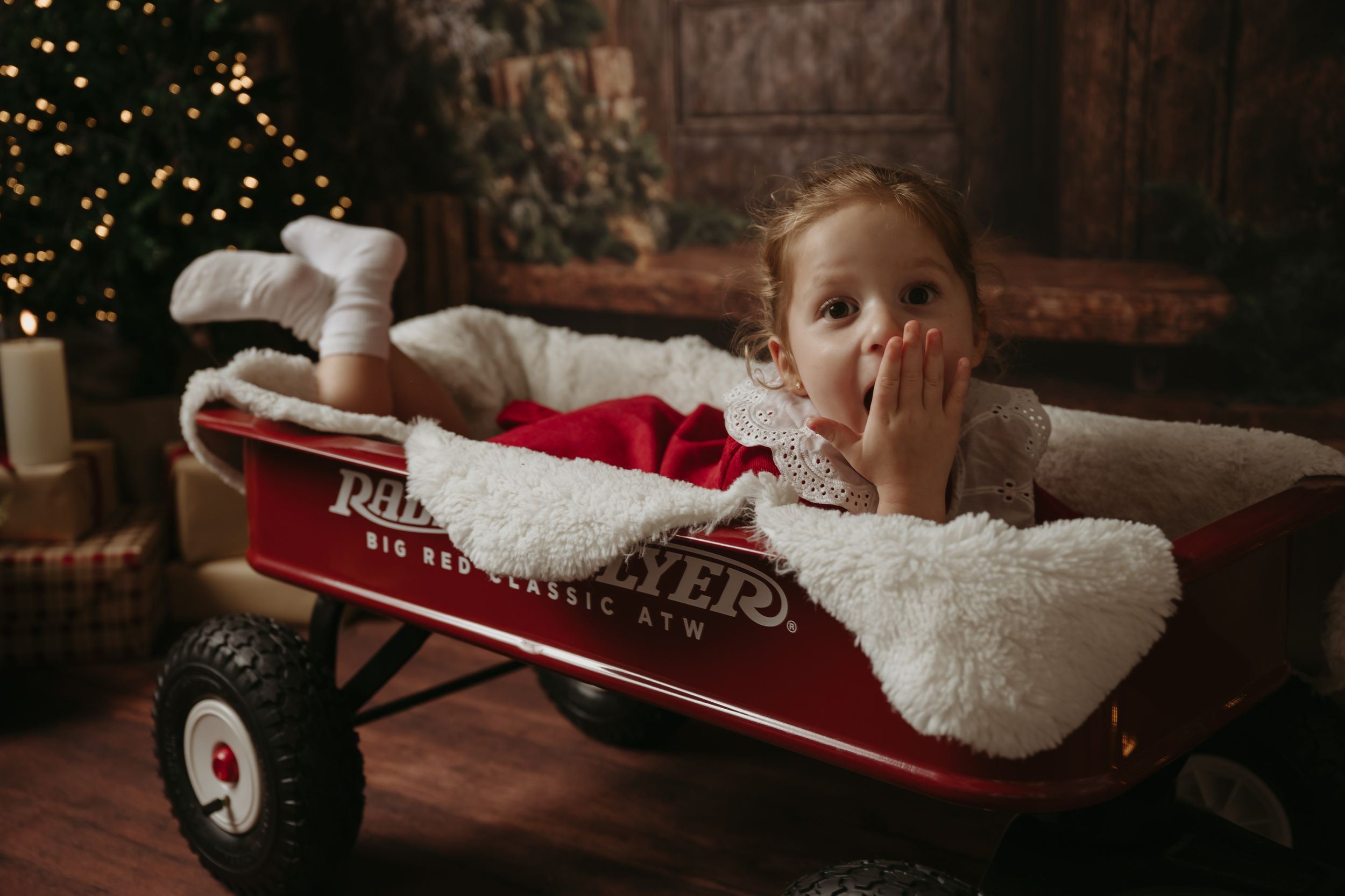 A child lies in a cozy red Radio Flyer wagon decorated for Christmas with a fuzzy white blanket and twinkling lights.