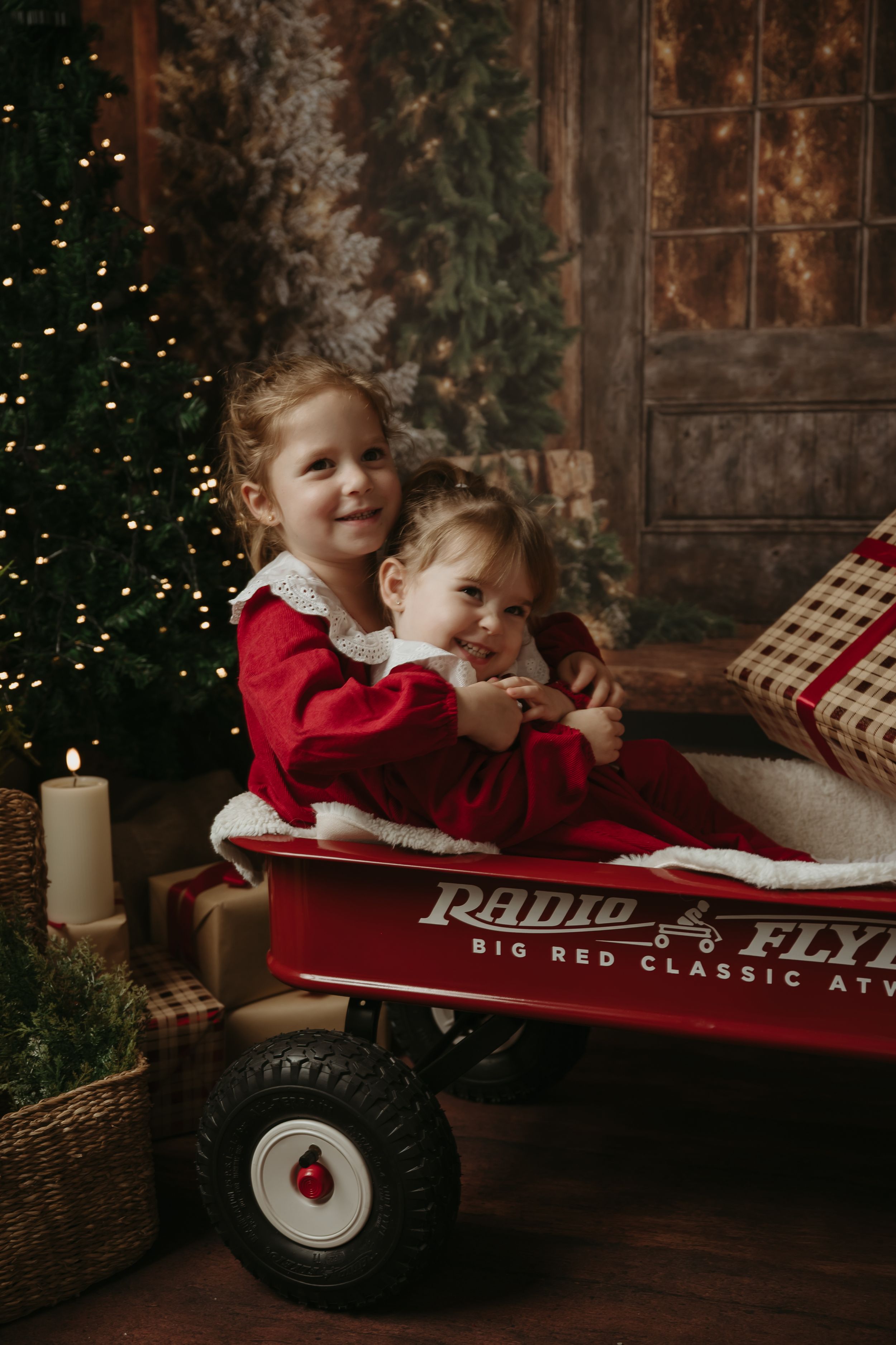 Two children in red outfits lay in a vintage red Radio Flyer wagon during a cozy Christmas photoshoot.