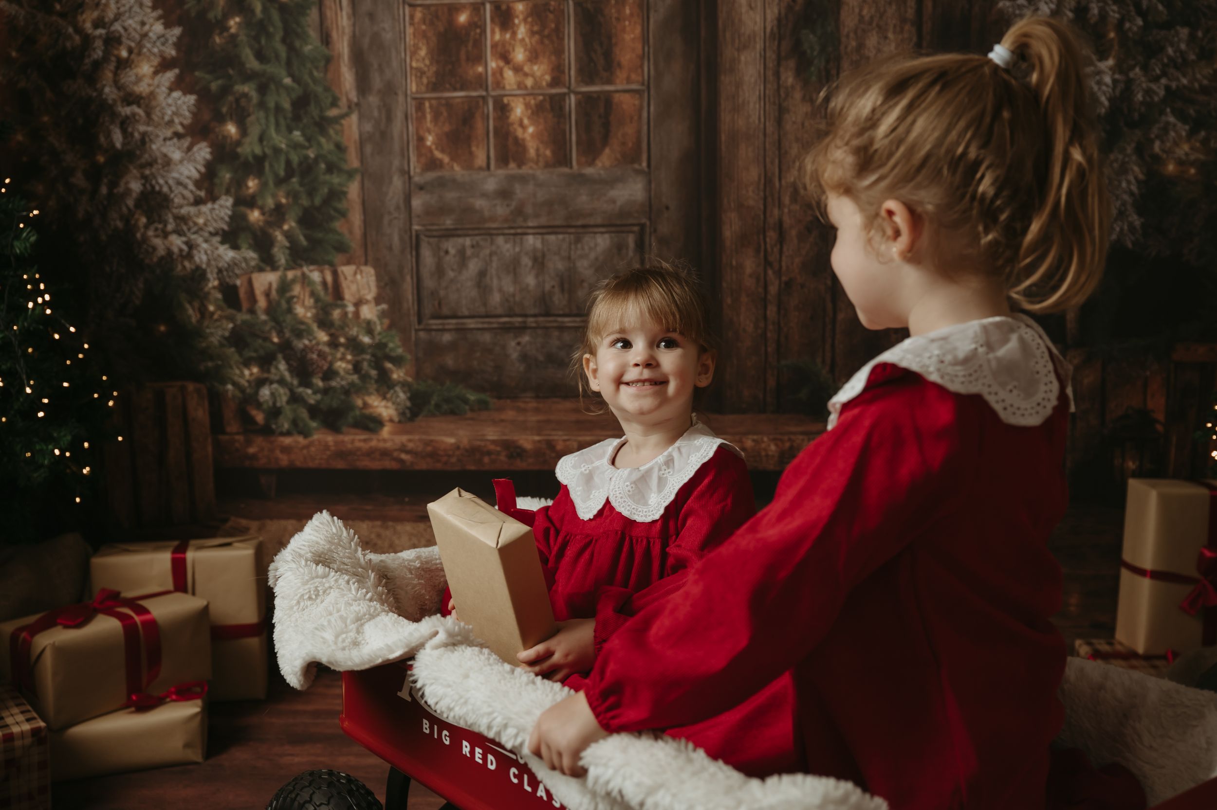 Two children in red holiday outfits sit together with wrapped Christmas presents and decorations in a cozy indoor setting.