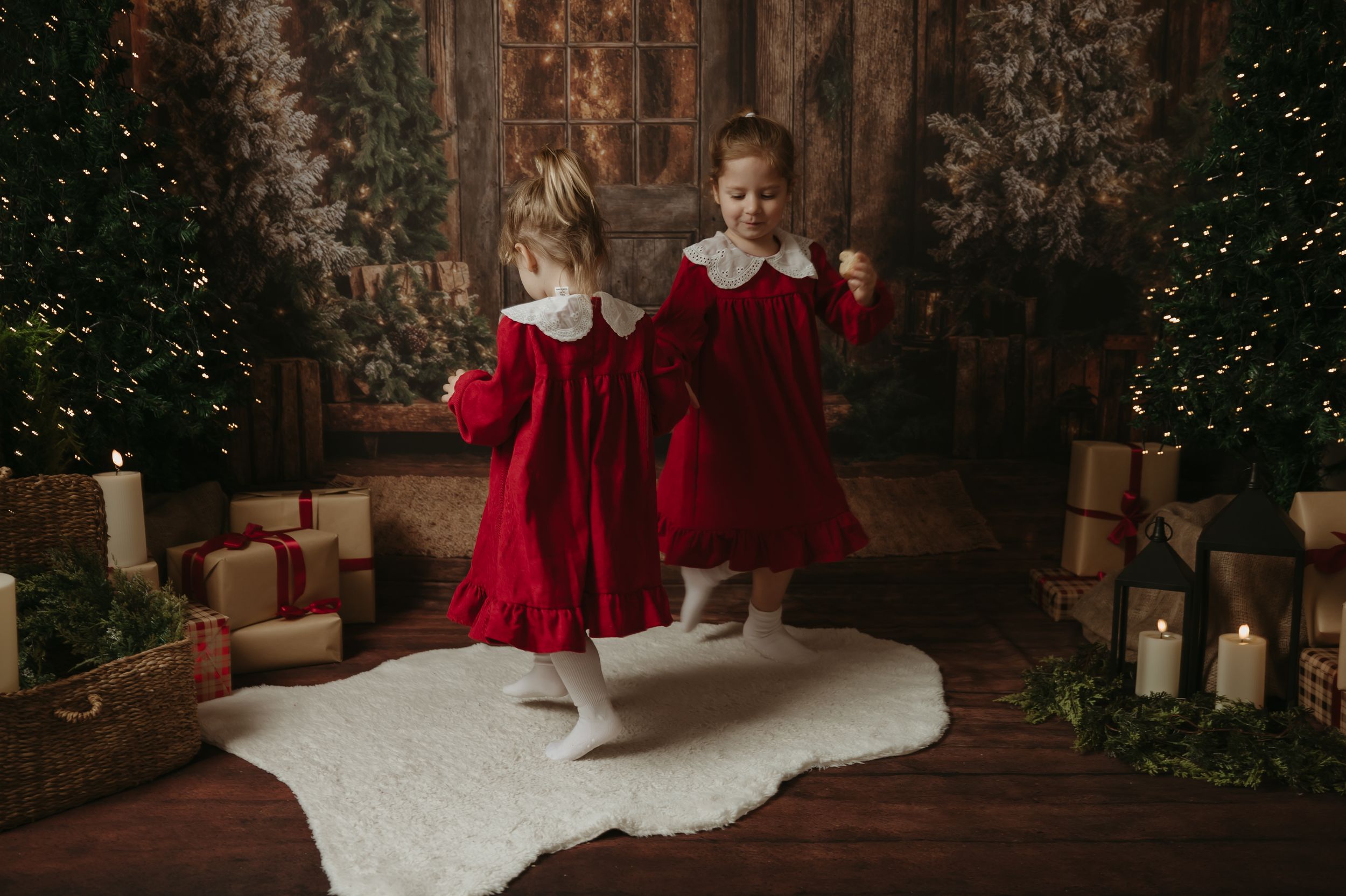 Children in red holiday dresses pose near Christmas decorations and presents in a rustic indoor setting.