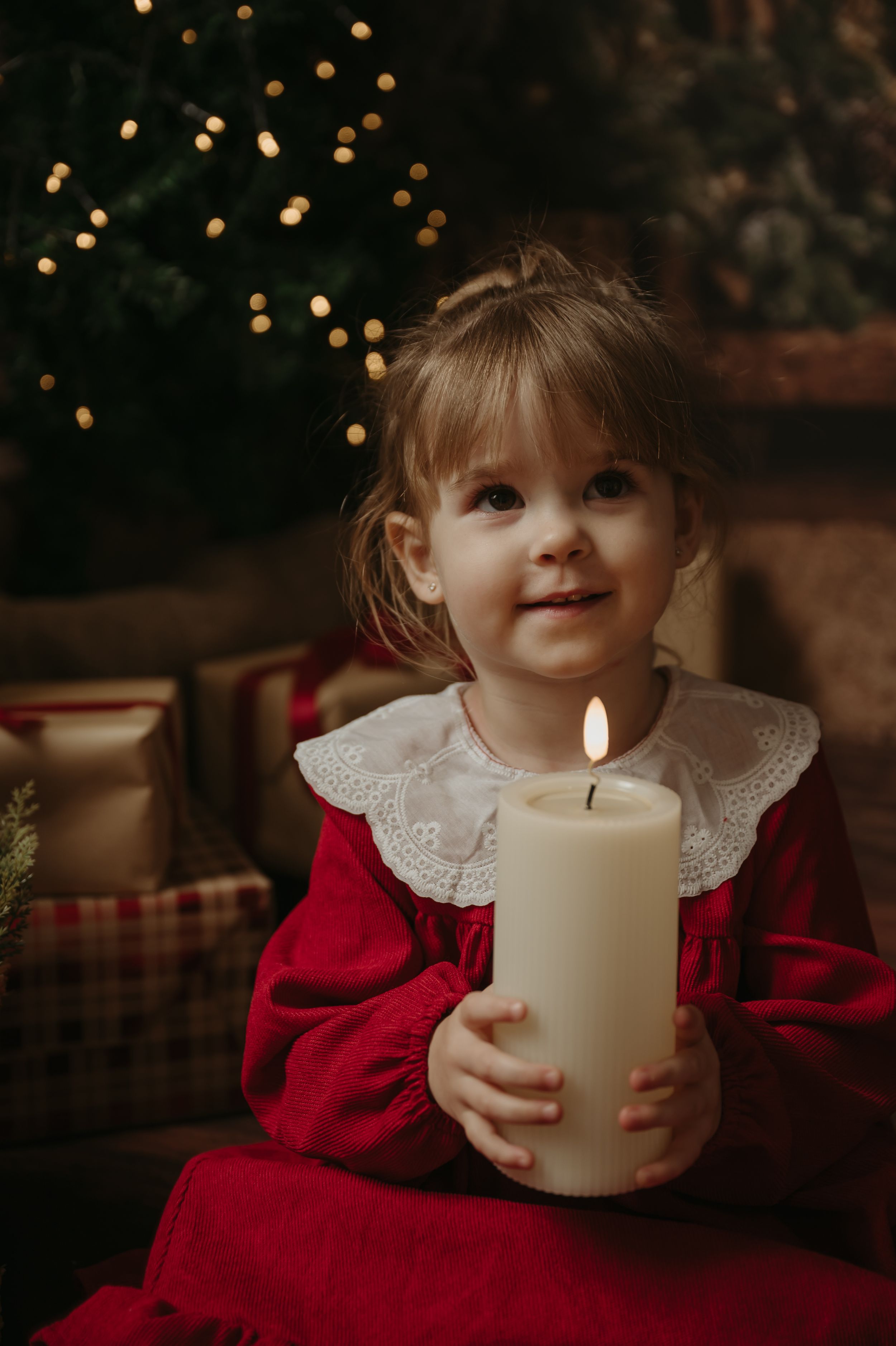 A young child in a red dress holds a white pillar candle while sitting in a cozy holiday setting with twinkling lights.