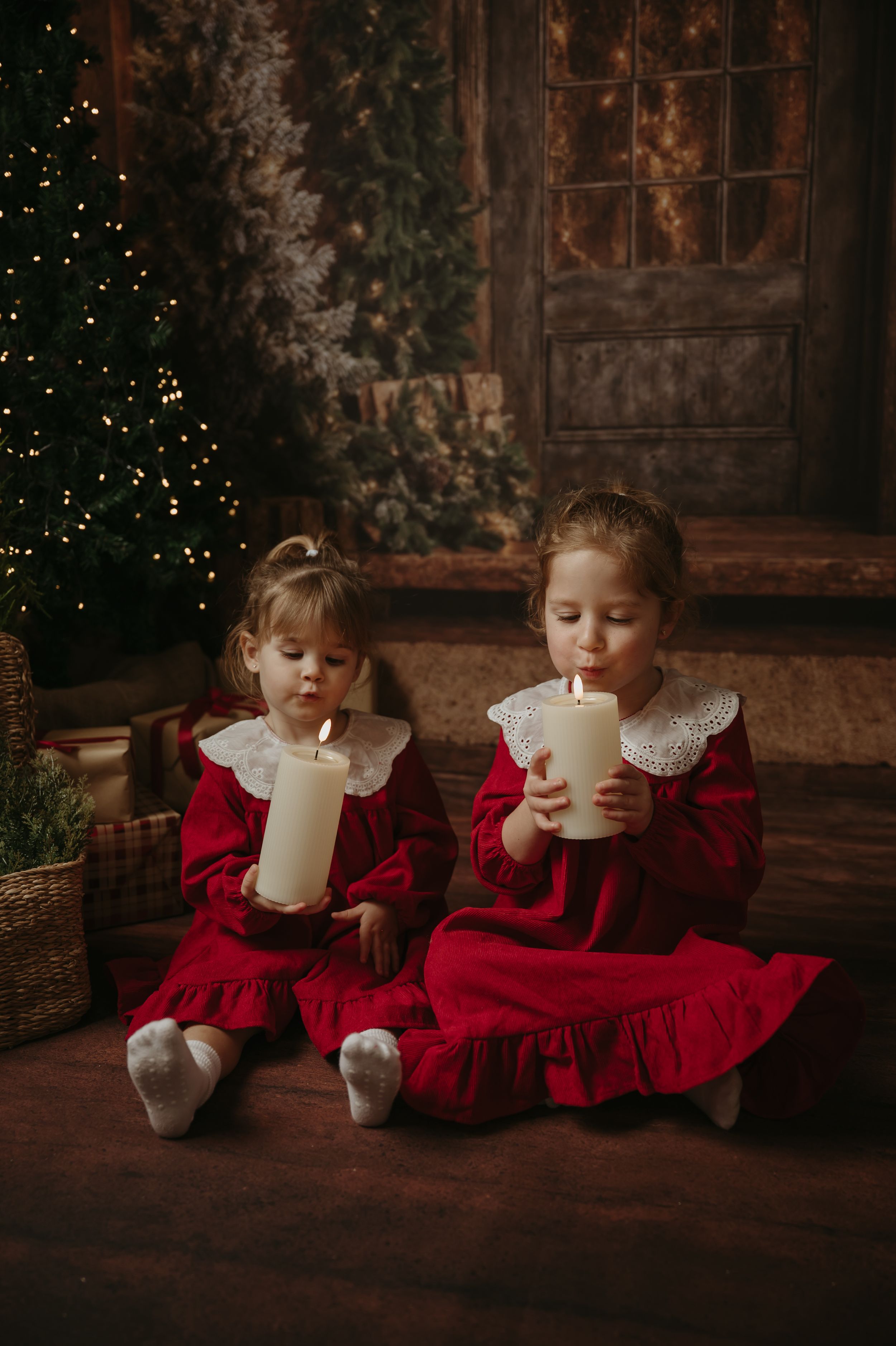 Two children in red holiday dresses sit together with large white candles in a cozy Christmas setting.